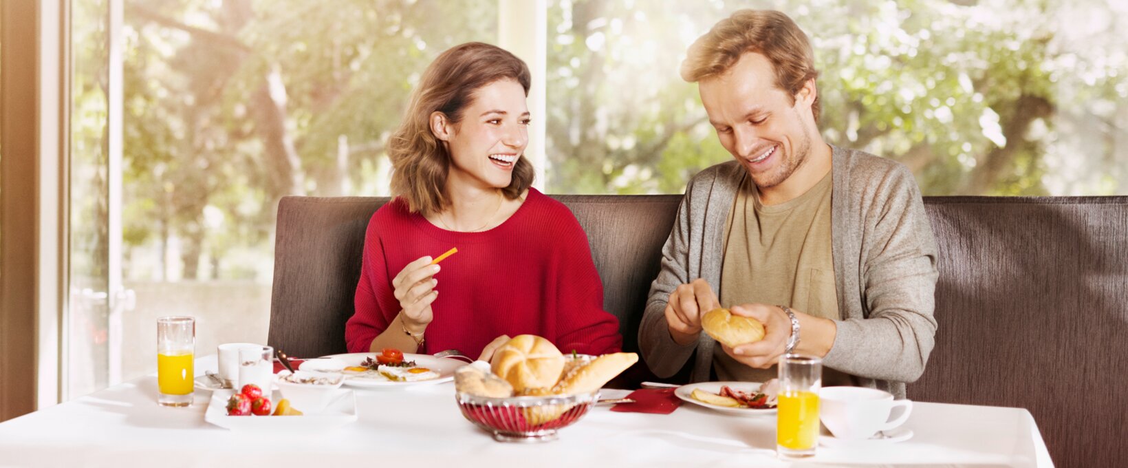 lachendes Paar am Frühstückstisch. Der Mann schneidet eine Semmel, während die Frau ihn anlächelt. 