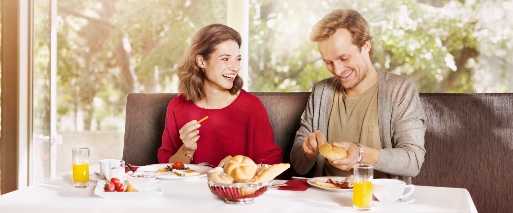 lachendes Paar am Frühstückstisch. Der Mann schneidet eine Semmel, während die Frau ihn anlächelt. 