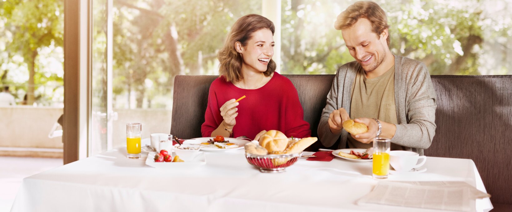 lachendes Paar am Frühstückstisch. Der Mann schneidet eine Semmel, während die Frau ihn anlächelt. 